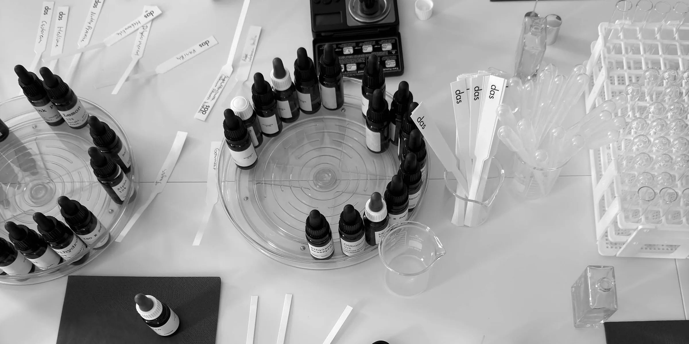 ein arbeitsplatz im workshop von oben: rohstoffflaschen auf drehtellern, eine feinwaage, beschriftete duftstreifen, pipetten und ein leerer flakon